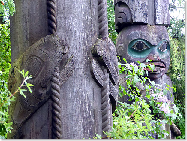 Hohe Kunst in altem Holz. Orkas an einem Seil knabbernd – Teil eines Totempfahls, der an einen Fischzug erinnert. Im Hintergrund ein weiterer Pfahl mit einer Maske.